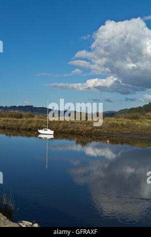 Small white sailboat anchored in calm reflective water under blue sky with clouds, estuary landscape. Stock Photo