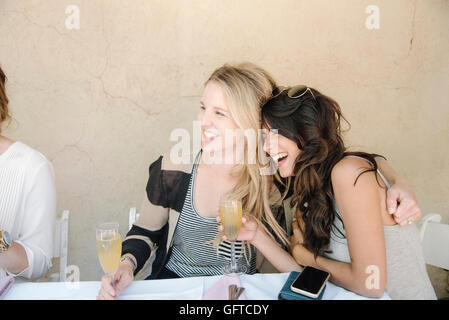 Two women sitting at a table hugging and laughing holding glasses of champagne Stock Photo