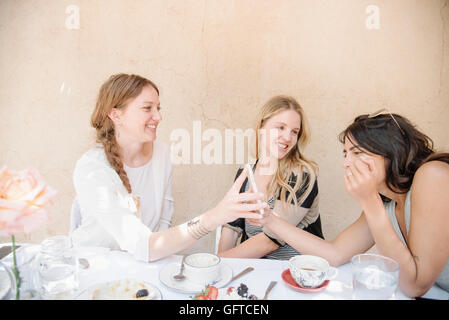 Three smiling young women sitting round a table with food and drink looking at a cell phone Stock Photo