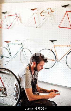 A man working in a bicycle repair shop sitting using his smart phone Stock Photo