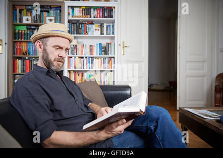 Man with beard reading a book in his living room on a leather couch Stock Photo