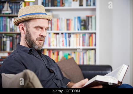 Student with hat sitting in front of a book shelf reading a book Stock Photo