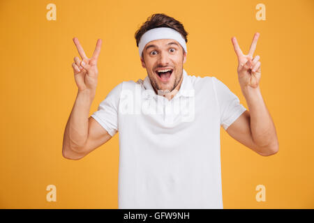 Happy excited young male athlete shouting and showing victory sign with both hands over yellow background Stock Photo