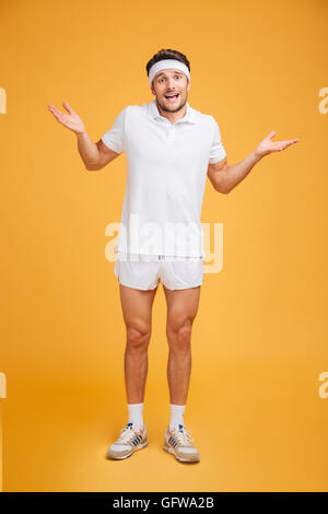 Full length of confused young sportsman standing and holding copyspace on palms over yellow background Stock Photo