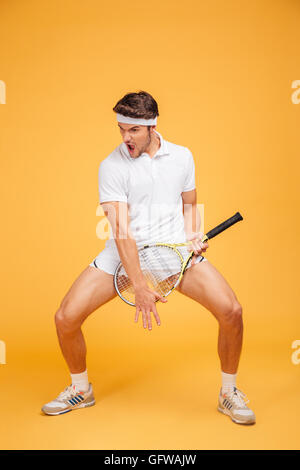 Funny playful young man tennis player with racket imitating playing guitar over yellow background Stock Photo
