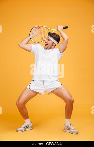 Amusing young man tennis player holding racket and having fun over yellow background Stock Photo