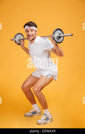 Cheerful young sportsman doing squats with barbell over yellow background Stock Photo