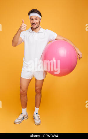 Young handsome sportsman holding fitness ball and showing thumb up isolated on the orange background Stock Photo