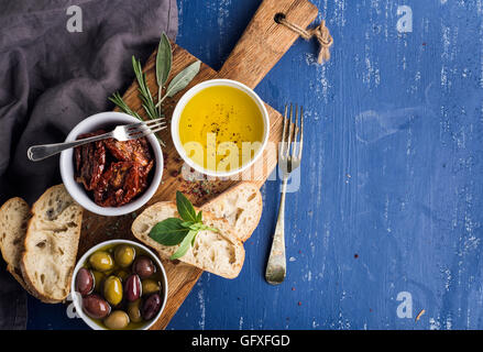 Mediterranean snacks set. Olives, oil, herbs and sliced ciabatta bread on yellow rustic oak board over painted dark blue background Stock Photo
