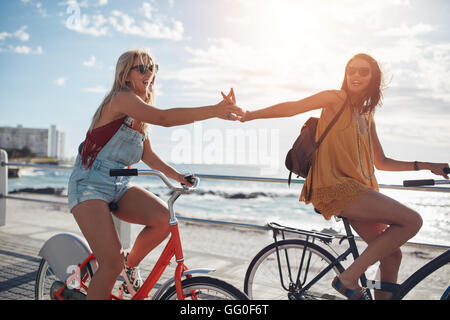 Female friends giving high five and riding bicycles along a seaside promenade on a summer day. Cheerful young women cycling outd Stock Photo