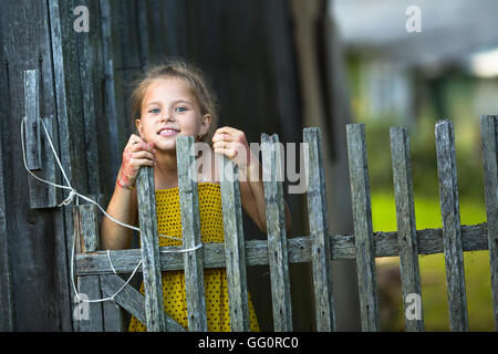 Cute little girl peeking out from behind the fence in the village. Stock Photo