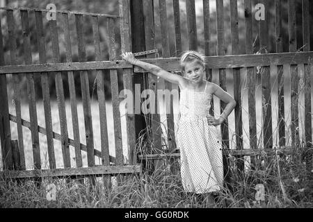 Cute little girl stands near a fence in the village. Black and white photo. Stock Photo