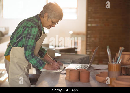 Female potter writing on a book while using laptop Stock Photo