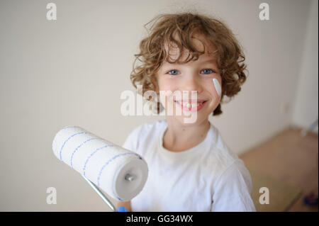 Cute little boy holding a roller for painting. His cheek was smeared with white paint. A boy with a cheerful smile, looking into Stock Photo