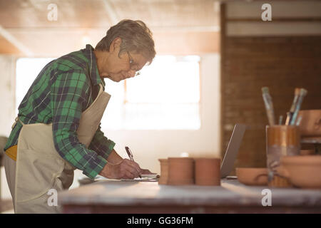 Female potter writing on a book while using laptop Stock Photo