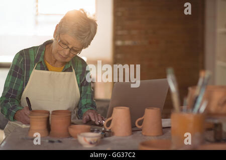 Female potter writing on a book while using laptop Stock Photo