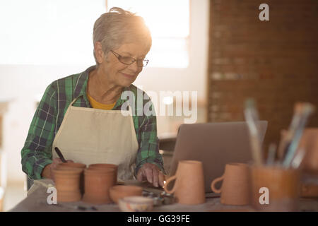 Female potter writing on a book while using laptop Stock Photo