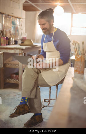Male potter sitting on stool using laptop Stock Photo