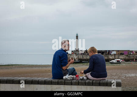 A man and a woman sitting on a wall on the seafront at Margate eating their lunch with the lighthouse in the distance Stock Photo