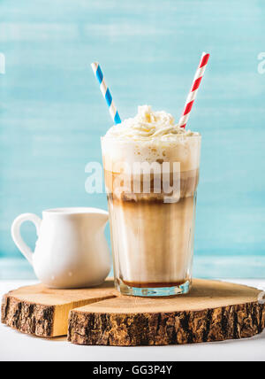 Latte macchiato with whipped cream in tall glass and pitcher on round wooden board over blue painted wall background Stock Photo