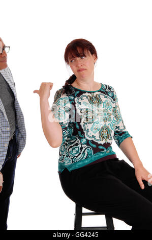 An upset woman sitting on a chair pointing back at her standing husband looking unhappy, isolated for white background. Stock Photo