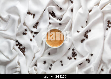 Coffee cup on a white silk fabric. Espresso and scattered beans. Top view. Stock Photo