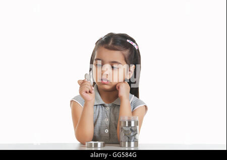 Cute girl holding Indian coin while sitting at table against white background Stock Photo