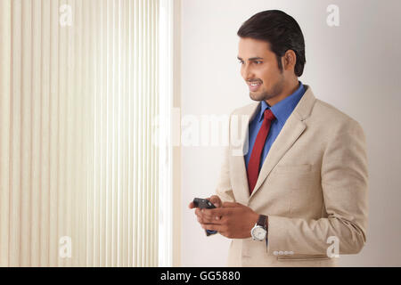 Young Indian businessman text messaging while looking through window blinds Stock Photo