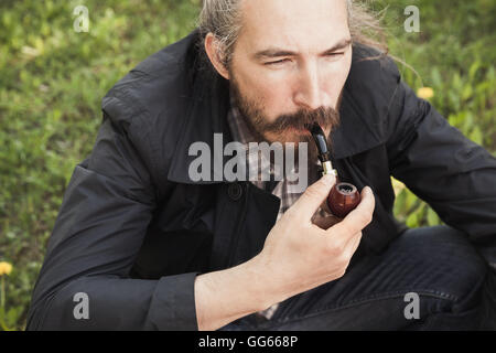 Asian man smoking a pipe on green grass in park, close-up portrait with selective focus Stock Photo