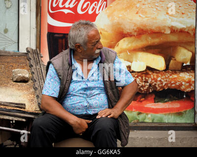 characterful large man with moustache and blue patterned shirt sits in front of fast food poster in street of Skopje Macedonia Stock Photo
