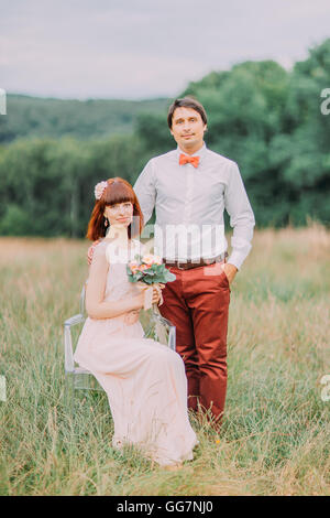 Beautiful bride outdoors in forest. She sits on a chair and keep wedding bouquet. Groom standing behind Stock Photo