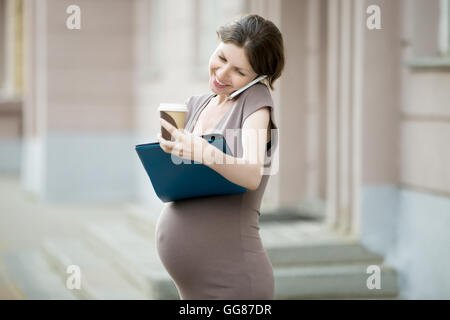 Active mom. Portrait of busy young pregnant business woman, talking on phone and making notes in the city. Smiling pregnant Stock Photo
