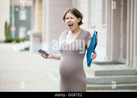 Portrait of unhappy young pregnant business woman walking in the city with document folder and mobile phone. Irritated office Stock Photo