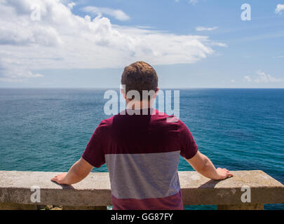 A young man looking out to a calm sea and flat horizon from a high vantage point at The Minack Theater in Cornwall, UK. Stock Photo