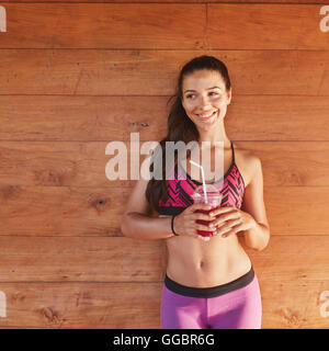 Portrait of healthy young woman with a glass of fruit juice after gym session standing against wooden background. Beautiful happ Stock Photo