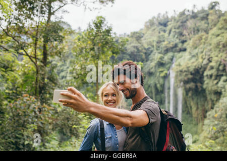 Happy young couple taking self portrait with their cell phone on hiking day. Couple taking picture in front of waterfall. Stock Photo