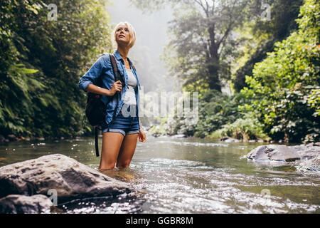 Outdoor shot of attractive young woman with backpack standing in a wilderness stream. Caucasian female hiker in creek water. Stock Photo