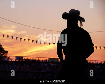 A silhouetted man in a cowboy hat stands in the bleachers watching a rodeo at sunset. Stock Photo