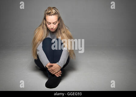 Sad young woman sitting in empty room on floor hands wrapped around legs looking down concept Stock Photo