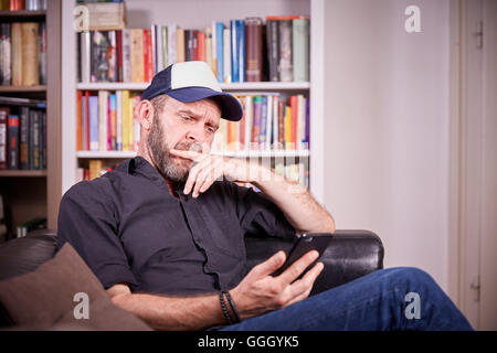 Man sitting on couch in living room looking on mobile phone brooding, pondering and looking seriously Stock Photo