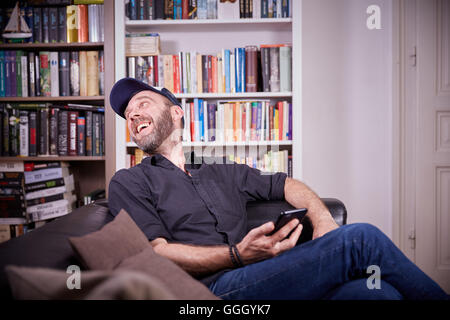 Man sitting on a couch in his living room with mobile phone laughing Stock Photo