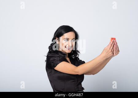 Smiling mid-30s woman in black blouse takes a cheeky selfie with her mobile phone. Studio image on white background with copy space to left Stock Photo