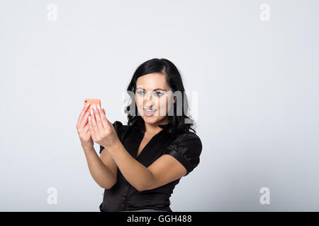 Smiling mid-30s woman in black blouse takes a cheeky selfie with her mobile phone. Studio image on white background with copy space to right Stock Photo