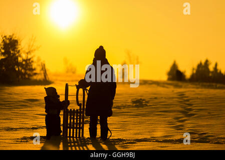 Mother standing with a small child and sled in the snow and looking at the winter sun, backlit, Aubing, Munich, Bavaria, Germany Stock Photo