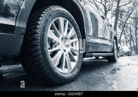 Fragment of black unidentifiable suv car, light alloy wheel with winter tires on dirty city road. Blue toned photo with selectiv Stock Photo