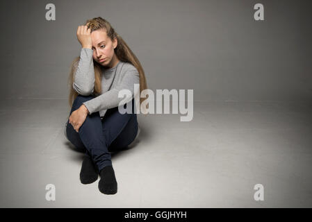 Sad young woman sitting in empty room on floor knees bent hand on leg other hand on head looking far away Stock Photo