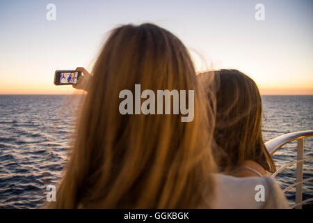 Two teenage girls taking a selfie photograph aboard cruise ship MS Deutschland (Reederei Peter Deilmann) at sunset, Atlantic Oce Stock Photo