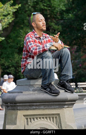 A young man pausing to meditate while sitting on a pillar and reading in Washington Square Park in New York City Stock Photo