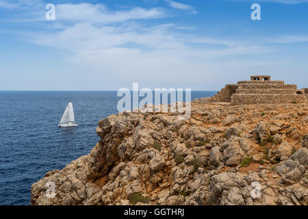 Boat sailing in front of a rocky cliff with an old stone Minorcan farm house above Stock Photo