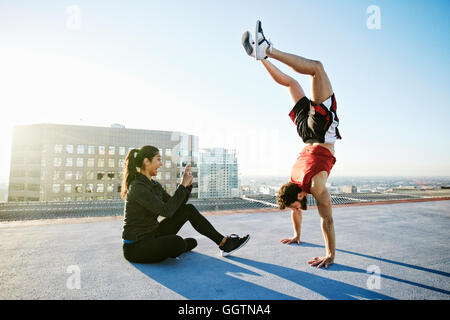 Woman photographing man doing handstand on urban rooftop Stock Photo